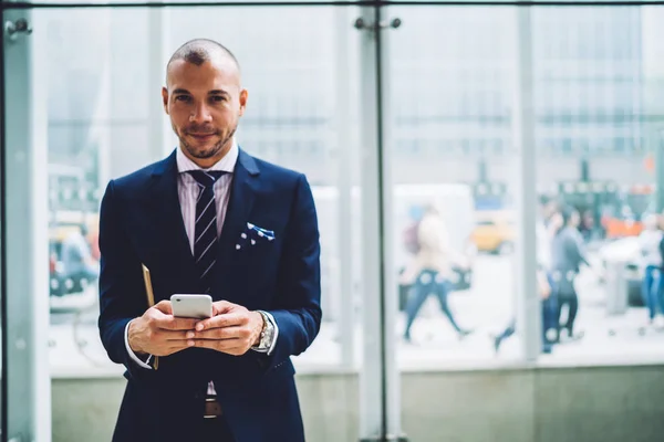 Proud CEO of corporation in formal wear banking via smartphone standing near copy space area for advertising, confident young businessman looking at camera checking notification on mobile phon