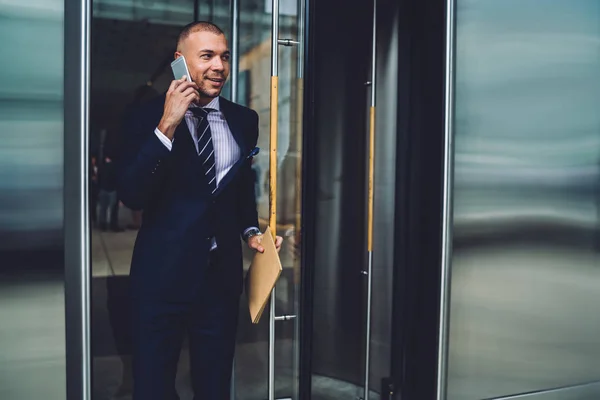 Smiling executive manager leaving building talking in phone with colleague about successful meeting, prosperous businessman in formal wear open doors sharing good news during phone conversation