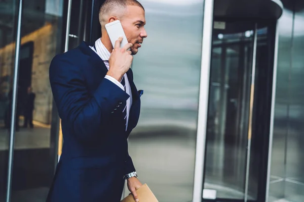 Serious businessman leaving building having phone conversation with operator checking money on banking account,confident corporate director talking on mobile hurrying for meeting getting out of office