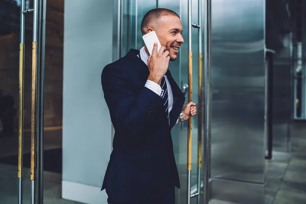 Cheerful financial manager sharing good news with colleague during phone conversation after successful meeting, positive male entrepreneur talking on mobile living office building after working day