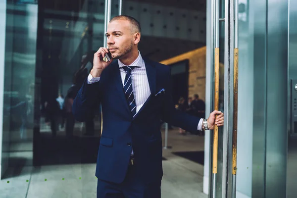 Confident male entrepreneur open door for leaving building after meeting making call to banking operator, handsome proud CEO talking on mobile with colleague standing in exit of office building