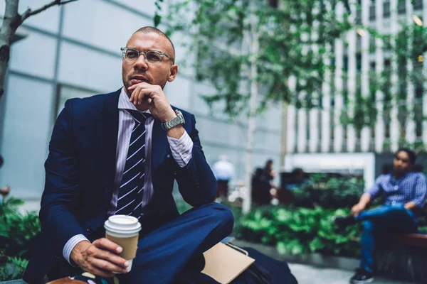 Thoughtful male entrepreneur in stylish eyeglasses pondering on working project during break outdoors, serious businessman holding coffee to go resting during free time on bench near green plants