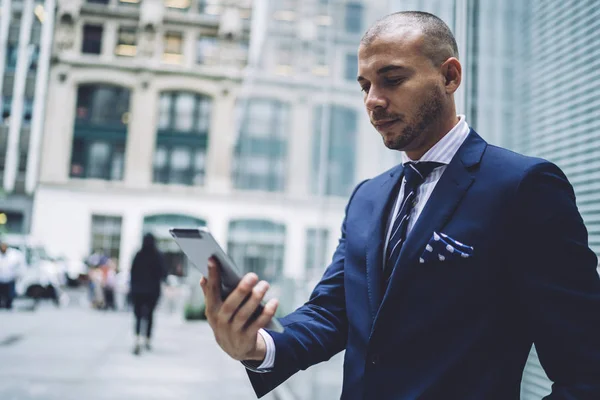 Pensive male entrepreneur in elegant wear making banking and paying for service online using tablet outdoors,prosperous trader checking news via application on portable pc standing near publicity area