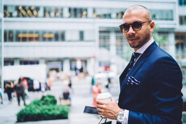 Portrait of  businessman in elegant outfit spending leisure outdoors with coffee to go standing on publicity area, serious boss in stylish sunglasses waiting for personal shopper looking at camer