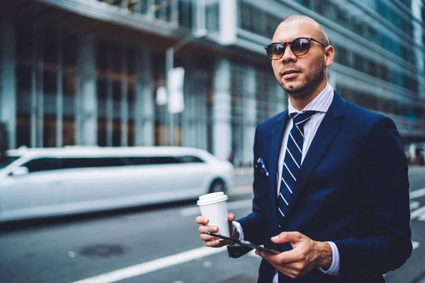 Self confident executive manager in trendy sunglasses standing on road with coffee takeaway waiting for cab, handsome businessman in formal wear using application on touchpad for calling taxi servic