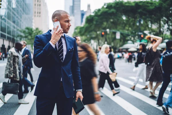 Confident male entrepreneur talking on phone standing on crowded crosswalk concentrated in conversation while everybody hurrying, serious businessman looking away choosing way while making mobile call