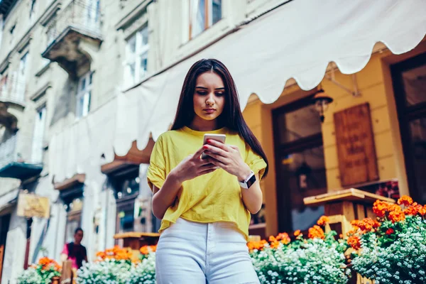 Pondering charming female blogger dressed in trendy outfit checking notification on smartphone connected to wireless internet while sharing and discussing ideas with followers in social network