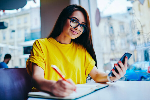 Портрет положительной брюнетки в модных очках, смотрящей в камеру при планировании рабочего графика с помощью приложения смартфона.
