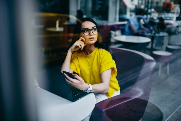 Contemplative charming graphic designer in trendy eyewear looking away and thinking about creative ideas for implementing in work.Gorgeous brunette student pondering while sitting in coffee shop