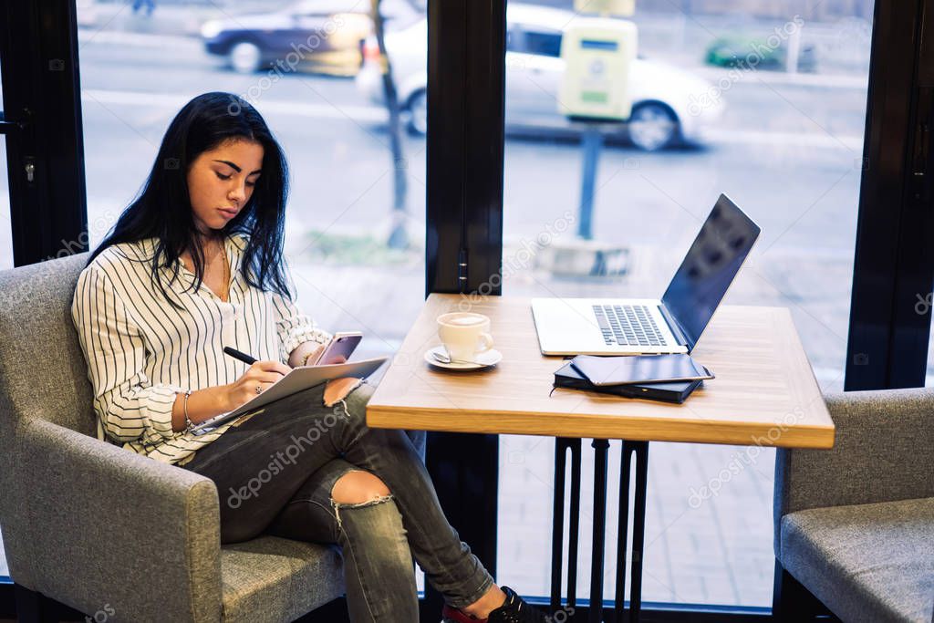 Young attractive businesswoman checking accounting from work papers while sitting in comfortable chair in coffee shop.Stylish pondering female student preparing for upcoming examination indoors