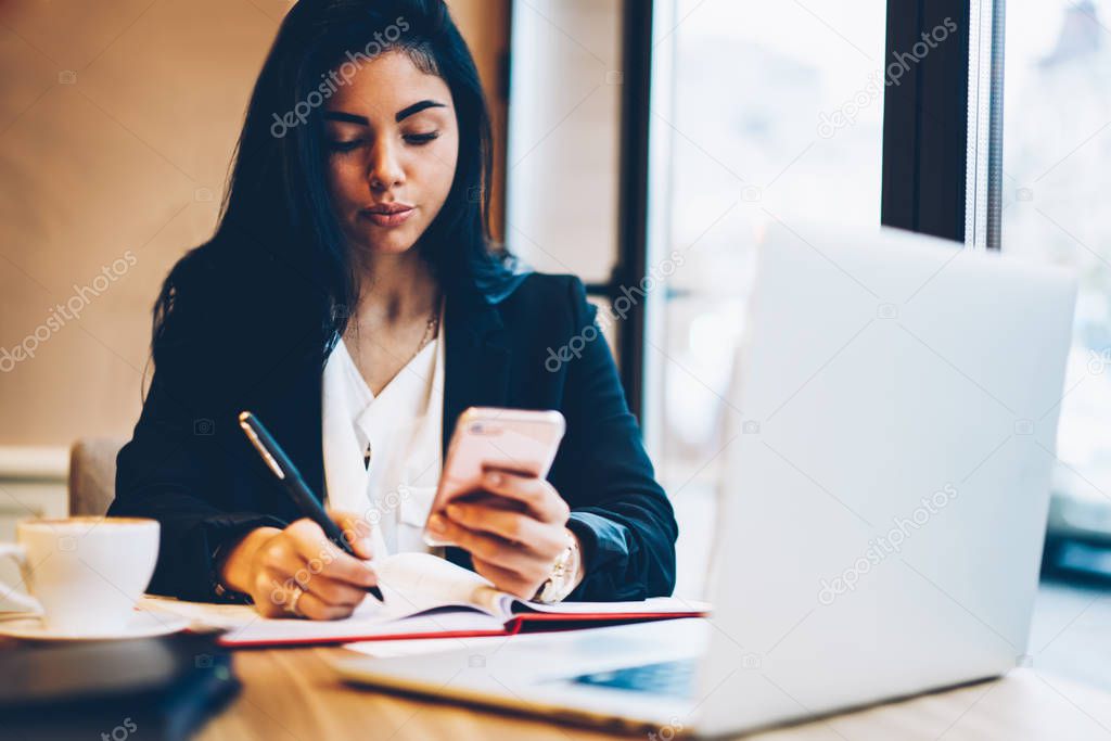 Charming student dressed in casual outfit making researches in websites on smartphone connected to 4G during preparing for coursework.Creative businesswoman recording to notepad ideas for startup