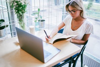 Metin içinde not etmek aşağı yazma ve ödev alan coworking içinde dizüstü aygıtı öğrenim sırasında şık gözlük yetenekli öğrenci. Yaratıcı tasarımcı gözlük kafede oturan kroki çizim