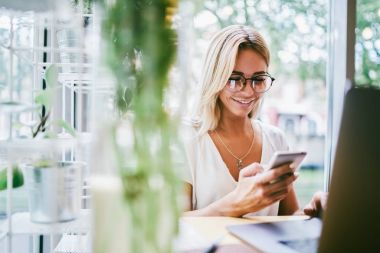 Optik Gözlük arkadaşlarıyla sohbet alanı coworking oturan modern telefon aracılığıyla ileti içinde pozitif kadın blogger. Smartphone cep telefonu ile komik yazı okuma gözlüğü neşeli genç kadın