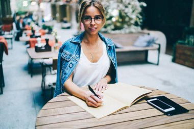 Şık gözlük kahvehane iç ahşap masada oturan yüzündeki gülümseme ile kameraya bakarken aşağı not defterinde kompozisyon yazma güzel genç hippi kız portresi yarım uzunluk