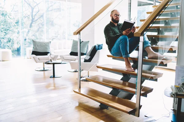 Young bearded guy fond of literature resting with favorite book at coy home interior, bearded man in spectacles reading  interesting bestseller sitting at stairs at modern designed apartments