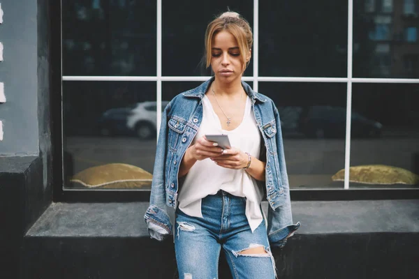时尚时髦的女孩穿着时髦的牛仔裤服装阅读短信收到的现代蜂窝 年轻妇女浏览互联网网页和做购物的数字智能手机在闲暇时间 — 图库照片