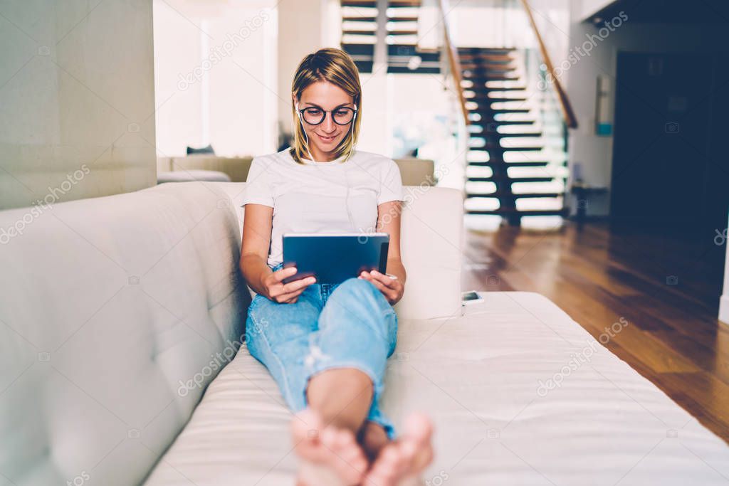 Positive young female in eyewear watching movie on tablet using earphones for entertaining during leisure at home interior, hipster girl reading news from networks and listening music from playlist