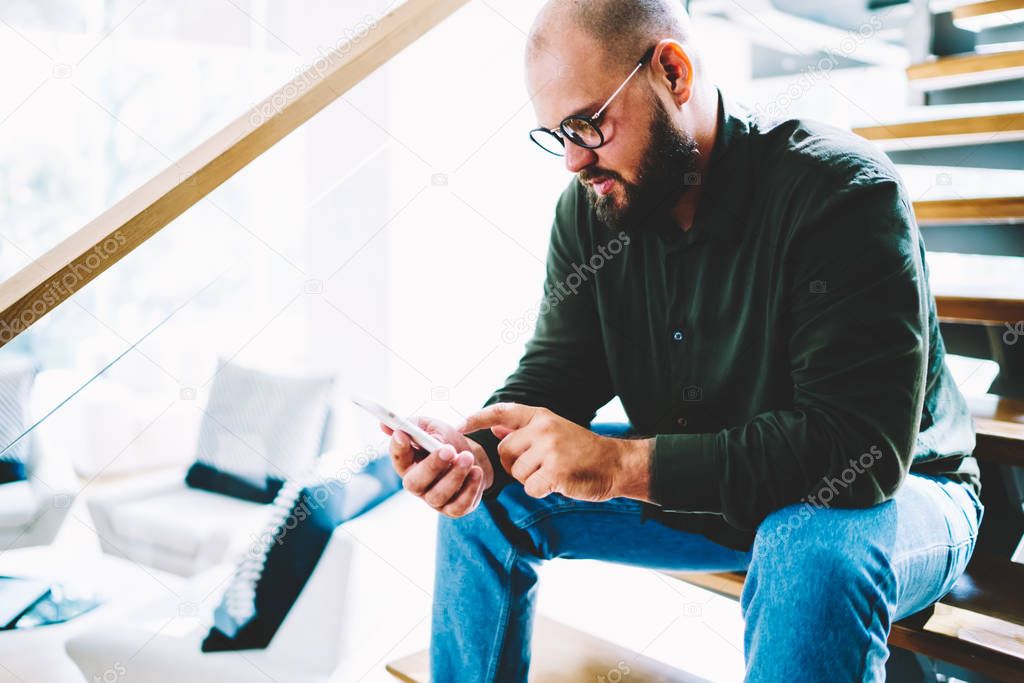 Pensive bearded man reading news from network sitting at stairs in modern designed apartment, hipster guy in eyeglasses checking banking account for making money transaction spending time at home