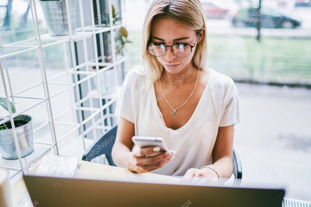 Top view of blonde young woman in optical stylish eyeglasses reading received sms message from friend sitting at table in coworking space.Female entrepreneur in spectacles chatting n social networks