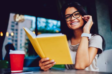 Neşeli esmer öğrenci serbest zaman sırasında ilginç bir kitap okuma edebiyat düşkün hippi kız en çok satan roman kafede kahve molası sırasında oturan arsa ile heyecanlı gülümseyen portresi
