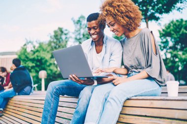 Başarılı afro Amerikalı arkadaşlarım şaka gülen ve modern cihaz bankta oturan kullanarak sırasında rahat giyim giymiş. Mutlu bir adam ve dizüstü bilgisayar üzerinde web sitesi komik video izlerken kadın