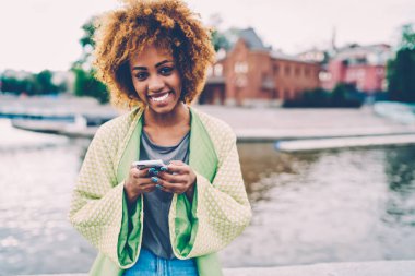 Yarım uzunlukta smartphone uygulama ile arkadaş komik iletiler göndermesi sırasında gülüyor kıvırcık saçlı oldukça neşeli kadın portresi. Mutlu traveler ağda eğleniyor sırasında kameraya bakıyor