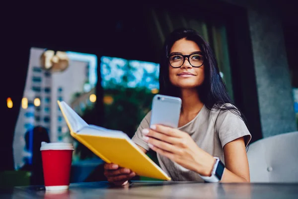 迷人的年轻女商人在眼镜检查每天的日程在乳制品和通知上的智能手机休息 有吸引力的学生阅读最喜欢的书在咖啡馆 而得到朋友的消息 — 图库照片