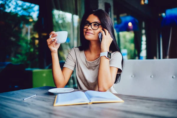 Positive female entrepreneur discussing work on phone with colleague during coffee break, attractive young woman in eyeglasses enjoying tea and conversation with friend on mobile in cafe interior