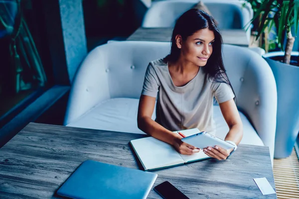 Thinking businesswoman writing notes while making research for project planning spending time in coffee shop, attractive student resting in cafe chatting in social networks via touchpad doing homework