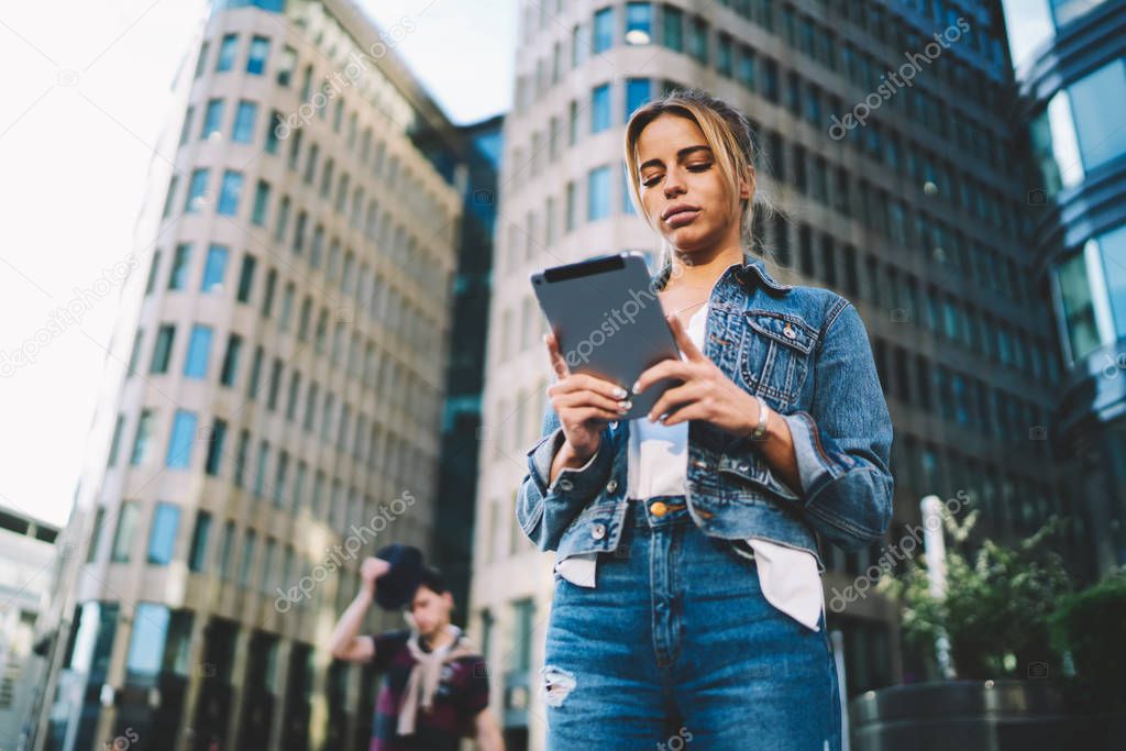Below view of pensive tourist looking at touch pad and searching right route on online navigator standing on street in city centre.Stylish young woman with modern tablet strolling in urban setting