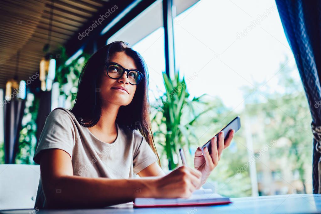 Serious businesswoman checking financial news on smartphone while making accountings in notebook,concentrated female entrepreneur sending files in networks waiting for feedback from colleague