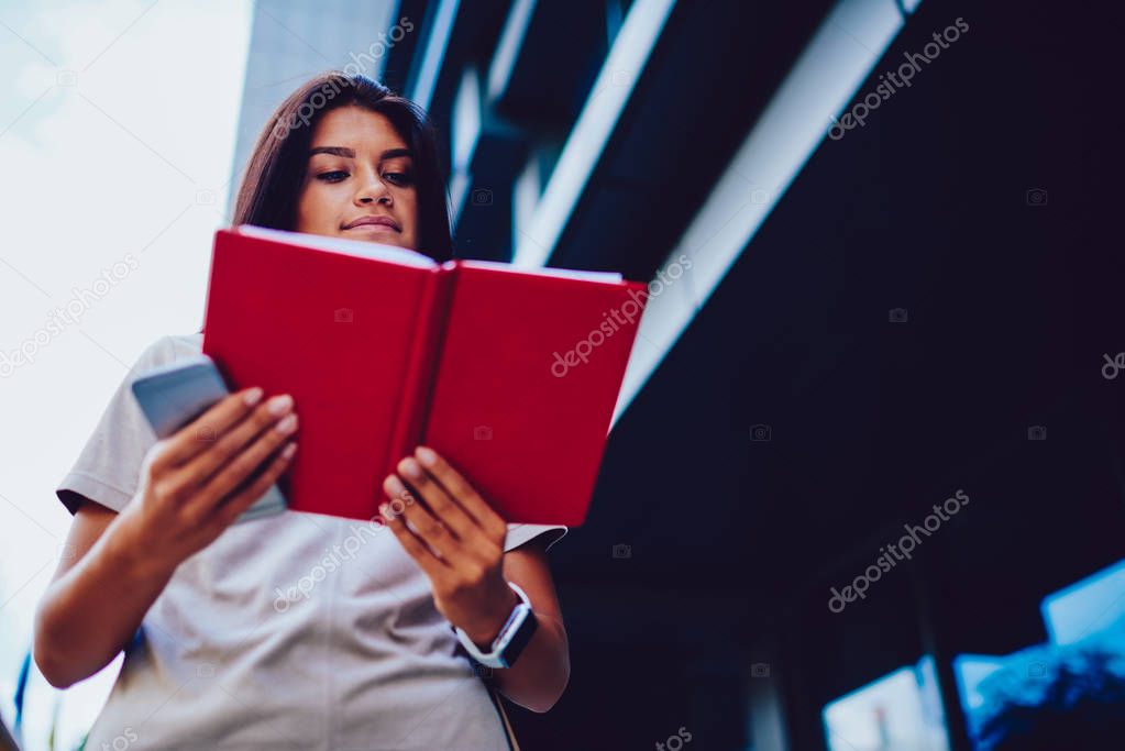 Concentrated brunette student reading book preparing for examination outdoors while walking, attractive hipster girl fond of literature enjoying interesting novel strolling in college campus
