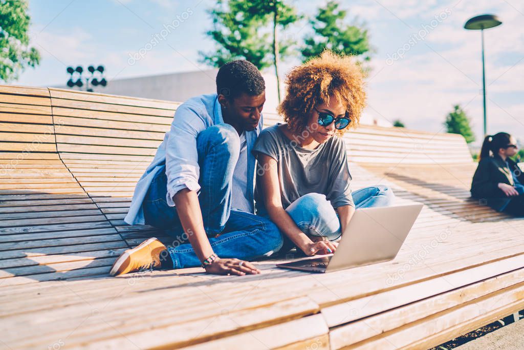 Attractive woman in sunglasses searching interesting movie for watching on laptop with male partner during resting at urban setting.Afro american couple using gadget while sitting on wooden bench