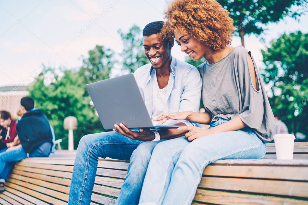 Successful afro american best friends dressed in casual clothing joking and laughing during using modern device sitting on bench.Happy man and woman watching funny video in website on laptop computer