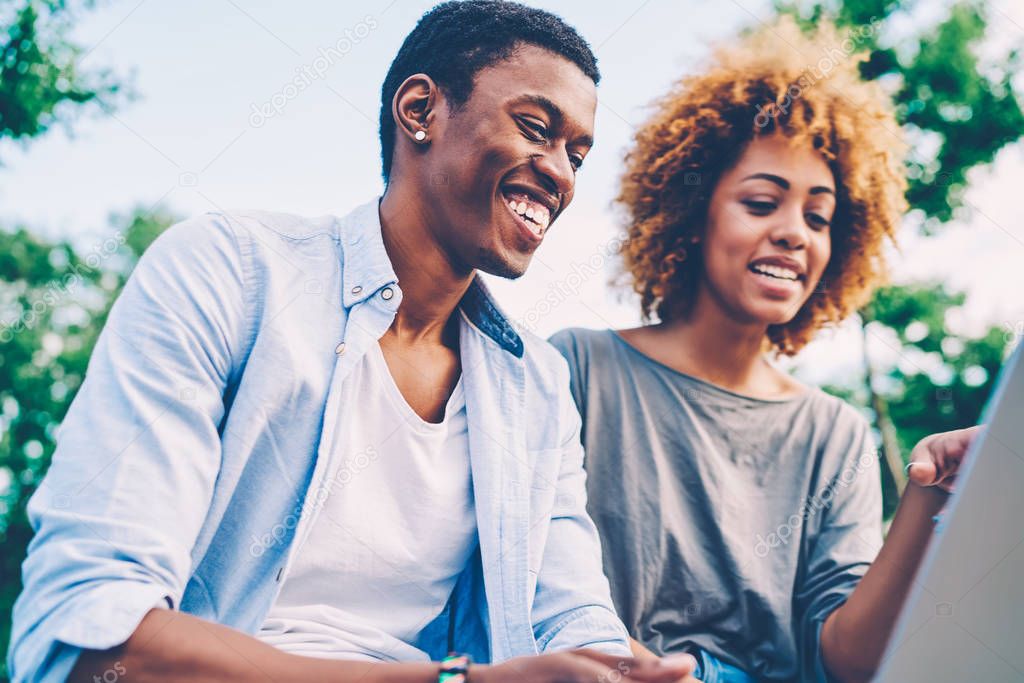 Cheerful male and attractive female laughing while watching funny TV show in webpage on laptop connected to wireless internet.Positive afro american hipsters enjoying free time at urban setting