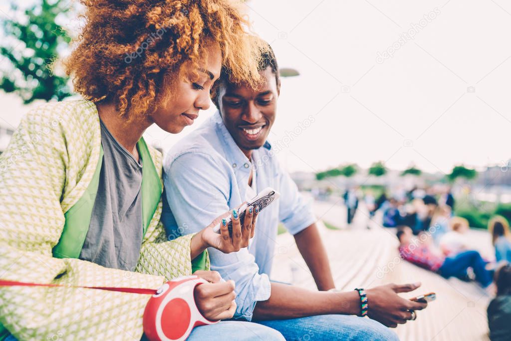 Cropped image of beautiful afro american woman showing interesting video in webpage on cellular for cheerful man sitting outside.Young happy couple recreating at urban setting with modern gadget