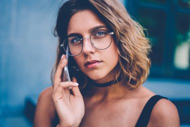 Uluslararası bağlantı kullanarak mobil konuşmayı düşünceli çekici hippi kız telefon görünümlü açık havada, vakit kameraya konuşurken gözlük ciddi genç kadın portresi