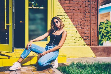 Çekici genç kadın içinde renkli kentsel ayarı arka plan açık havada playlist smartphone cep telefonu ile zevk kamera poz arayan rahat kıyafet giymiş hippi kız üzerinde oturan kulaklıklar müzik dinleme