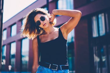 Şık giyimli hippi kız ücretsiz zaman açık havada sırasında güneşli bir gün, güzel genç kadın kentsel ayarları arka sokakta durup poz fotoğraf makinesi bakarak zevk güneş gözlüğü portresi