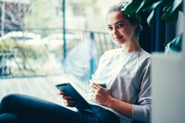 Yarım uzunluk portre rekreasyon lezzetli içecek ve dokunmatik yüzey ile kapalı oturma sırasında zevk çekici öğrenci. Zamana Cafe kopya alanı ile pencere yakın harcama genç hippi kız