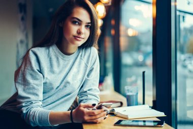 Yarım uzunluk portre yetenekli öğrencinin ders projesi için telefon üzerinde farklı web sitelerinde bilgi aramak. Kadın yazarı yazılım smartphone cep telefonu ile güncelleştirilirken kameraya bakıyor