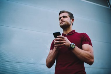 Aşağıda görünümü yakışıklı hipster adam uzakta kısa mesaj reklam için gri duvarının yakınında yürüyüş sırasında dijital cep telefonunda sms arkadaşlar yazarken arıyorum. Genç adam elinde modern smartphone ile
