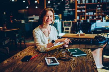 Neşeli işkadını mola Cafe iç çalışma günü planlama portre fikirlerle, çekici Bayan uzak iş için ekipman ve ileti örneği ile masada oturan kamera bakarak memnun.