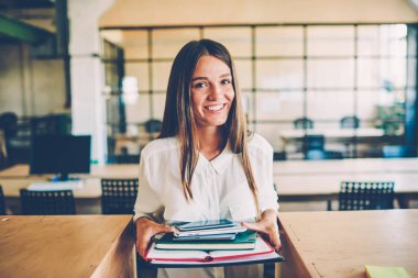 Varoluş kitap ve Kırtasiye sınıfta, mutlu genç esmer kadın öğrenme zevk alan coworking içinde kamera ayakta bakarak vakit holding neşeli teen öğrenci portresi