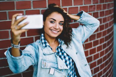 Muhteşem kıvırcık saçlı poz ve selfie resimleri ile kendi web sitesi açık havada duvar promosyon arka plan karşı ayakta yüklemek için yeni fotoğraf uygulaması teyel çekici genç kadın