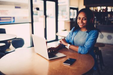 Neşeli afro Amerikan öğrenci dizüstü cihaz otururken kameraya gülümseyen bağlı kablosuz 4 g için portresi internet ve çalma listesinden Kulaklığımda yeni şarkıları ile en sevdiğiniz müzik dinleme