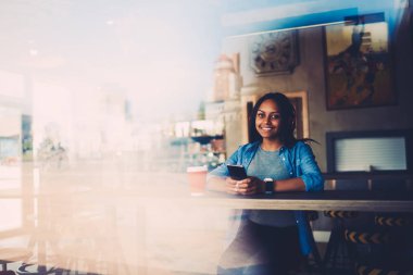 Afro Amerikan kadın blogger dinleme ses kulaklık girişi portresi kafede oturan kamera bakarak telefon aygıtı bağlı. Reklam alanı için reklam metin iletiniz