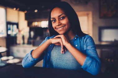 Kameraya bakarak ve serbest zaman zevk kahve dükkanında oturmuş gülümseyen güzel afro Amerikalı genç kadın yarım uzunluk portre. Gündelik kıyafeti giymiş siyah deri ile çekici hippi kız