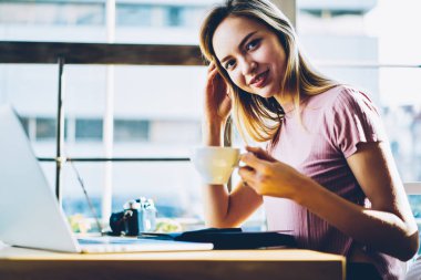 Neşeli kafeterya sahibi lezzetli kapuçino dizüstü bilgisayar çalışma için 4 g bağlıyken el kameraya bakarak fincan ile yarım uzunluk portre internet ve çevrimiçi belgeleri kontrol