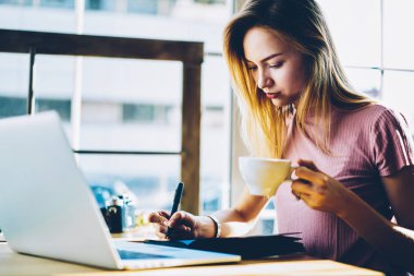 Genç girişimci iş süreci lezzetli kapuçino kapalı dizüstü bilgisayar başında oturan zevk kendi Not Defteri'nde düzenlemek için planı yazıyor. Öğrenci el günlüğü kayıtları belirterek kahve ile